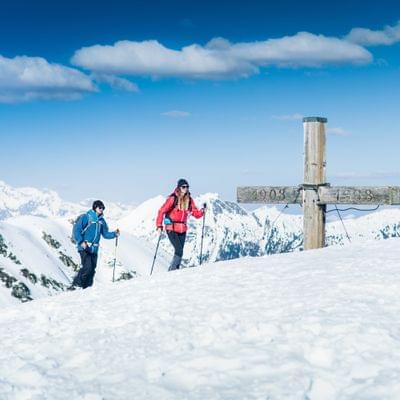 Two hikers trekking through snowy alpine landscape near summit cross